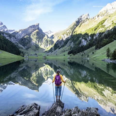 Eine Person mit Rucksack und Wanderstöcken steht auf einem Felsen neben einem ruhigen See, umgeben von grünen Hügeln und felsigen Bergen. Das klare Wasser spiegelt die Landschaft und den hellen Himmel wider und schafft eine malerische Landschaft.