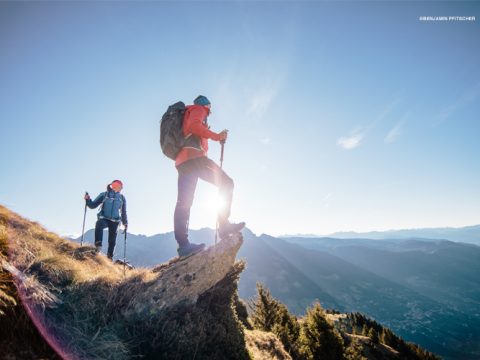 Zwei Wanderer stehen auf einem felsigen Gipfel und genießen das Sonnenlicht. Einer steht mit Wanderstöcken am Rand, der andere in der Nähe. Sie blicken auf eine weite Landschaft aus Bergen und Tälern unter einem klaren blauen Himmel.