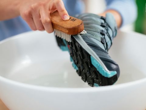 Eine Person reinigt einen blau-grauen Sportschuh mit einer Holzbürste über einem mit Wasser gefüllten weißen Becken. Der Fokus liegt auf der Schuhsohle und der Bürste, die mit ihr in Kontakt kommt.
