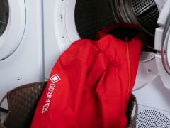 Eine leuchtend rote Gore-Tex-Jacke liegt in einer offenen Waschmaschine, ein Teil davon hängt über der Tür. In der Nähe steht ein brauner geflochtener Korb, der unten links teilweise sichtbar ist.