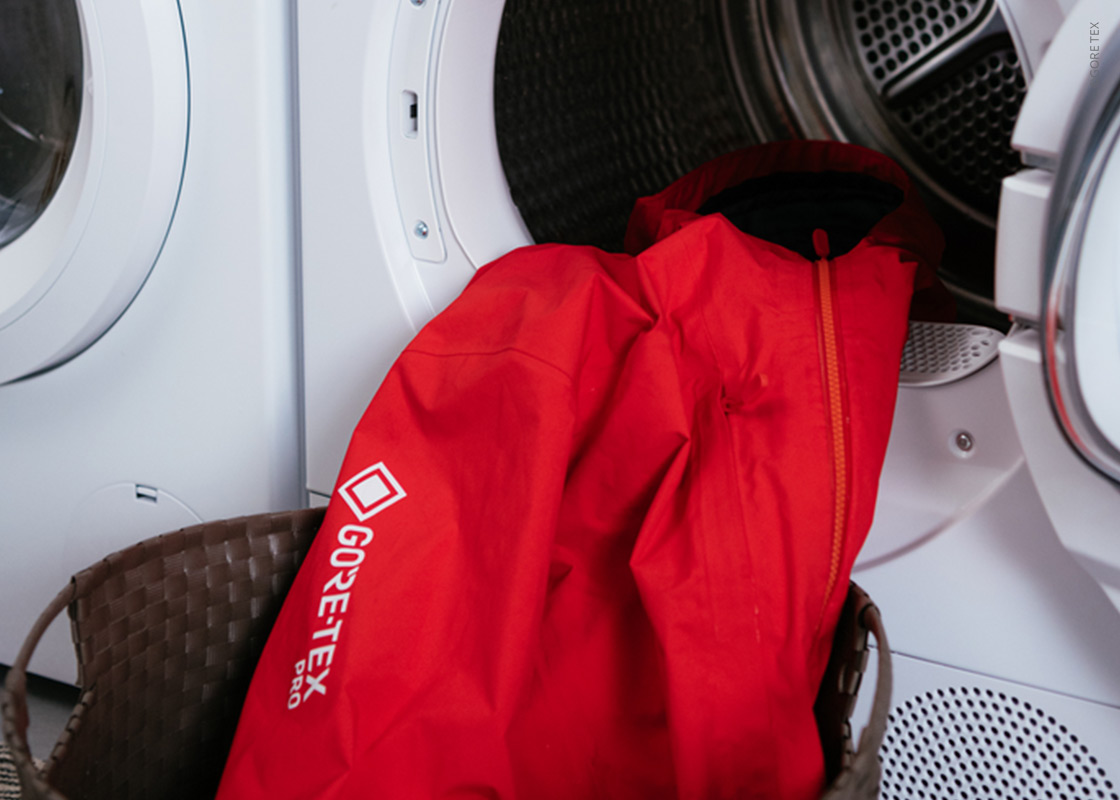 Eine leuchtend rote Gore-Tex-Jacke liegt in einer offenen Waschmaschine, ein Teil davon hängt über der Tür. In der Nähe steht ein brauner geflochtener Korb, der unten links teilweise sichtbar ist.