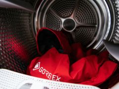 Rote Gore-Tex-Jacke in einer Waschmaschinentrommel, aus der Nähe von der Türöffnung aus betrachtet. Die Jacke ist zusammengeknüllt und zeigt das Gore-Tex-Logo. Das metallische Innere der Trommel ist deutlich zu erkennen.