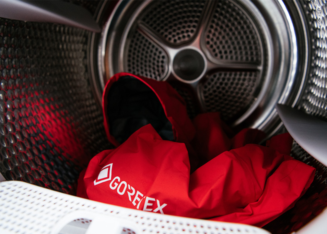 Rote Gore-Tex-Jacke in einer Waschmaschinentrommel, aus der Nähe von der Türöffnung aus betrachtet. Die Jacke ist zusammengeknüllt und zeigt das Gore-Tex-Logo. Das metallische Innere der Trommel ist deutlich zu erkennen.