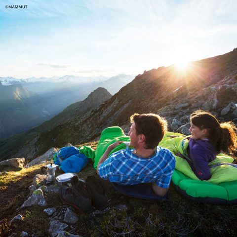 Ein Mann und eine Frau liegen in Schlafsäcken auf Isomatten und blicken auf die Berge bei Sonnenaufgang. Campingausrüstung, darunter Tassen und ein Rucksack, liegt in der Nähe. Der Himmel ist klar und wirft einen warmen Schein auf die Szene.