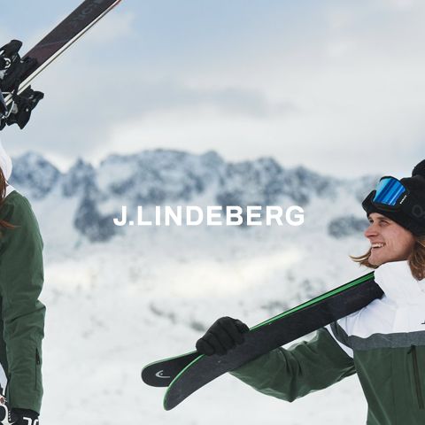 Zwei Personen in Skiausrüstung stehen in einer verschneiten Berglandschaft und halten jeweils Skier auf ihren Schultern. Sie lächeln sich an. In der Mitte ist das Logo „J. Lindeberg“ zu sehen.