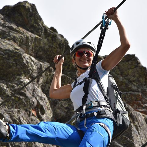 Eine Person mit Helm, Sonnenbrille und Kletterausrüstung steigt mithilfe eines Seils einen felsigen Berg hinauf. Sie lächelt, trägt ein weißes Hemd und blaue Hosen. Der Himmel ist klar und im Hintergrund ist felsiges Gelände zu sehen.