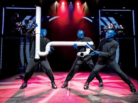 Drei blau bemalte und schwarz gekleidete Darsteller halten ein großes weißes Rohr in einer industriellen Bühnenkulisse mit bunter Beleuchtung. Sie stehen auf der Bühne vor einem dynamischen Hintergrund mit roten und blauen Lichtern und einer darüber liegenden Bandsilhouette.