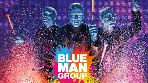 Drei Figuren mit blau bemalten Gesichtern treten energisch vor einem lebendigen, farbenfrohen Hintergrund auf. Sie halten Trommelstöcke, die von Farbspritzern umgeben sind. Die Worte „Blue Man Group“ sind deutlich zu sehen.