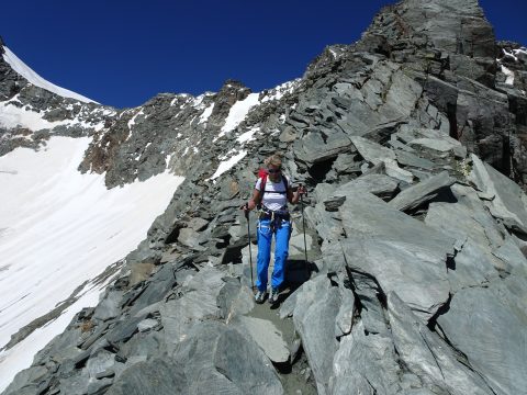 Eine Person in weißem Hemd und blauer Hose wandert mit Wanderstöcken einen felsigen Bergpfad mit Schneeflecken hinauf. Der Himmel ist klar und blau und im Hintergrund sind schroffe Gipfel zu sehen.