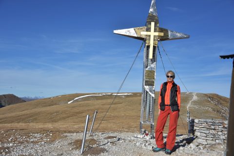 Eine Person in roter Outdoor-Kleidung steht neben einem großen Metallkreuz auf einem felsigen Hügel. Der Himmel ist klar und die umliegende Landschaft ist karg mit sanften Hügeln im Hintergrund.