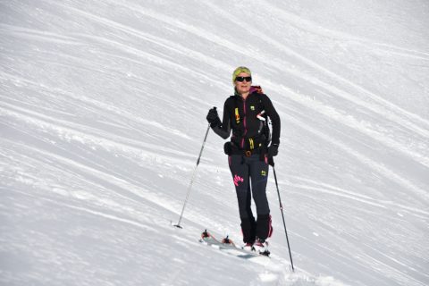 Eine Person in Wintersportkleidung fährt einen verschneiten Hang hinunter. Sie trägt einen Helm, eine Sonnenbrille und hat Skistöcke dabei. Der Hintergrund ist eine helle und gleichmäßige Schneefläche.