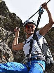 Eine Person mit Helm, Sonnenbrille und Kletterausrüstung lächelt beim Klettern. Sie hält sich an einem Seil fest, im Hintergrund ist ein Fels zu sehen. Die Person trägt ein weißes Hemd und eine blaue Hose.