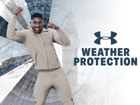 Eine Person in beiger Sportbekleidung posiert energisch vor einem schneebedeckten, bergähnlichen Hintergrund mit einem Uhrenturm. Das Under Armour-Logo und der Text „Weather Protection“ sind auf der rechten Seite deutlich zu sehen.
