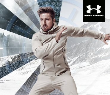Ein Mann in einem beigen Under Armour-Trainingsanzug steht in sportlicher Pose vor einem dynamischen Hintergrund mit Schnee und Bergen. Das Under Armour-Logo befindet sich in der oberen rechten Ecke.
