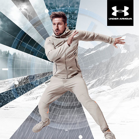 Eine Person in einem beigen Under Armour-Trainingsanzug posiert athletisch vor einem dynamischen Hintergrund aus schneebedeckten Bergen und geometrischen Mustern. In der oberen rechten Ecke ist das Under Armour-Logo zu sehen.