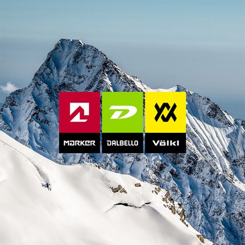Ein schneebedeckter Berggipfel unter klarem Himmel, mit drei überlappenden farbigen Quadraten in der Mitte, die jeweils ein Logo zeigen: Rot mit weißem „A“ für Marker, Grün mit weißem „D“ für Dalbello und Gelb mit schwarzem „V“ für Völkl.