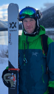 Eine Person in einer grün-blauen Skijacke steht da und hält ein Paar Skier. Auf ihrem Helm trägt sie eine reflektierende Schutzbrille. Im Hintergrund sind schneebedeckte Berge und ein See zu sehen.