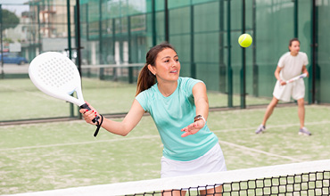 Eine Frau in einem hellblauen Hemd spielt Padel, hält einen Schläger und konzentriert sich auf einen Ball in der Luft. Ein weiterer Spieler steht im Hintergrund auf einem Außenplatz.