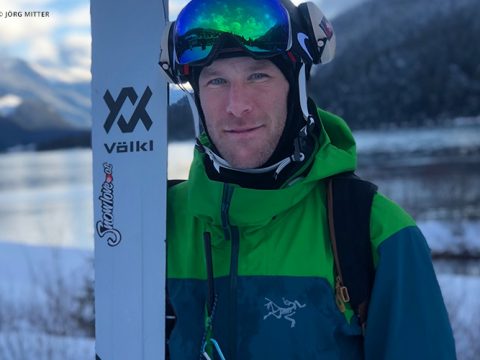 Eine Person in einer grün-blauen Jacke hält Skier und steht im Freien mit schneebedeckten Bergen und einem See im Hintergrund. Sie trägt reflektierende Skibrillen und Ohrenschützer.