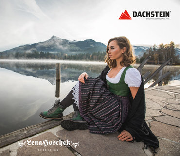 Eine Frau in traditioneller Kleidung sitzt auf einem Steinweg am Ufer eines Sees. Sie blickt auf das ruhige Wasser, umgeben von Bergen und Bäumen. Der Himmel ist bedeckt. In den Ecken sind die Logos von Lena Hoschek und Dachstein zu sehen.