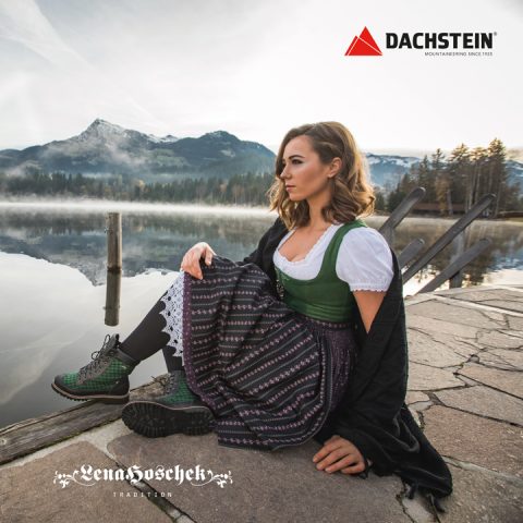 Eine Person in traditioneller Kleidung sitzt auf einem Steg am Seeufer mit Bergen und Nebel im Hintergrund. Sie trägt Wanderschuhe und ein gemustertes Kleid. Logos für „Lena Hoschek Tradition“ und „Dachstein“ sind sichtbar.