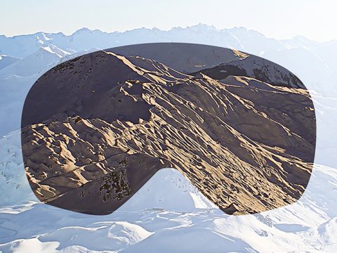 Eine schneebedeckte Berglandschaft wird durch die Silhouette einer Skibrille betrachtet. Die Berggipfel sind sonnenbeschienen, und die Schatten heben das zerklüftete Gelände hervor. Im Hintergrund ist ein klarer Himmel zu sehen.