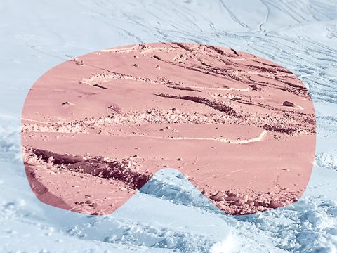 Eine verschneite Landschaft wird durch eine rosa getönte Schutzbrille betrachtet. Der Schnee außerhalb der Schutzbrille erscheint weiß, während er innerhalb der Schutzbrille einen rosa Farbton aufweist und Spuren und unebenes Gelände zeigt.