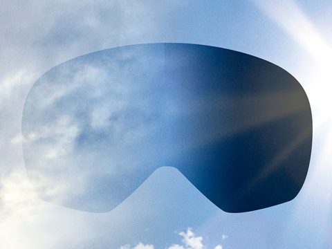 Die Linse einer Skibrille schwebt vor einem strahlend blauen Himmel mit vereinzelten Wolken. Sonnenlicht erzeugt auf der rechten Seite des Bildes einen Linsenreflexionseffekt.