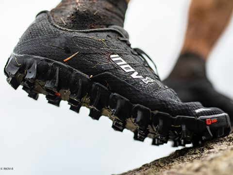 Nahaufnahme des Fußes einer Person, die einen schwarzen Trailrunning-Schuh mit robuster Sohle und der Aufschrift „INOV8“ trägt und auf einen felsigen Untergrund tritt. Der Schuh hat ein strukturiertes Design und scheint schlammig zu sein, was auf eine Aktivität im Freien hindeutet.