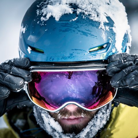 Nahaufnahme einer Person, die einen blauen Helm und eine reflektierende Skibrille trägt und diese mit beiden Händen zurechtrückt. Schnee bedeckt Helm und Brille, während die Person eine gelbe Jacke trägt. In der Spiegelung ist eine verschneite Landschaft zu sehen.