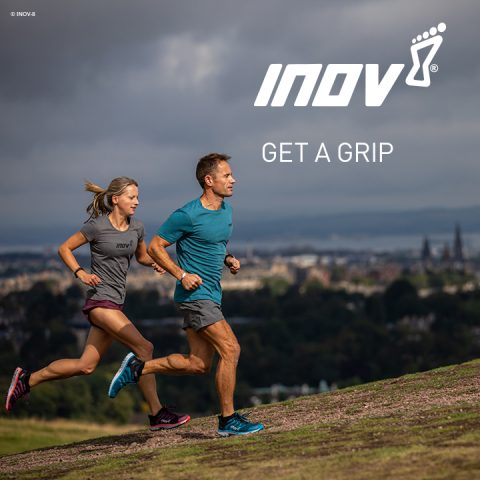 Ein Mann und eine Frau laufen einen grasbewachsenen Pfad bergauf, im Hintergrund ist eine Stadtlandschaft zu sehen. Beide tragen Sportkleidung. Der Himmel ist bewölkt. Oben steht „INOV8 GET A GRIP“ mit einem Logo.