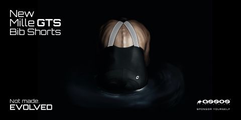 Eine Person ist von hinten in Radlerhosen zu sehen, in der oberen linken Ecke steht der Text „New Mille GTS Bib Shorts“. Das Bild hat einen dunklen Hintergrund, der die reflektierenden Träger und das Material der Hose hervorhebt.