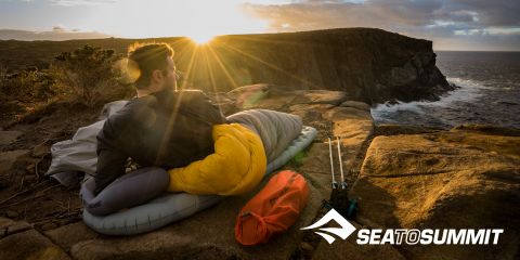 Eine Person liegt auf einer Isomatte, eingewickelt in einen gelben Schlafsack, und beobachtet einen Sonnenaufgang über einer Küstenklippe. Campingausrüstung liegt neben ihr, und die Meereswellen brechen gegen die Felsen. Das Logo „SEATOSUMMIT“ befindet sich in der unteren rechten Ecke.