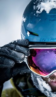 Nahaufnahme einer Person mit blauem Helm und reflektierender Skibrille, die mit einer behandschuhten Hand die Brille zurechtrückt. Auf Helm und Kleidung liegt Schnee, das Gesicht ist teilweise sichtbar. Die Spiegelung in der Brille zeigt eine verschneite Landschaft.