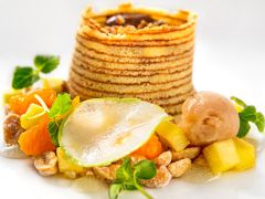 Ein Gourmet-Dessert besteht aus einem hohen, spiralförmigen Gebäckturm gefüllt mit Nüssen, umgeben von Obstwürfeln, einer dünnen Birnenscheibe, Grünzeug und einer Kugel Sorbet, alles elegant auf einem weißen Teller präsentiert.
