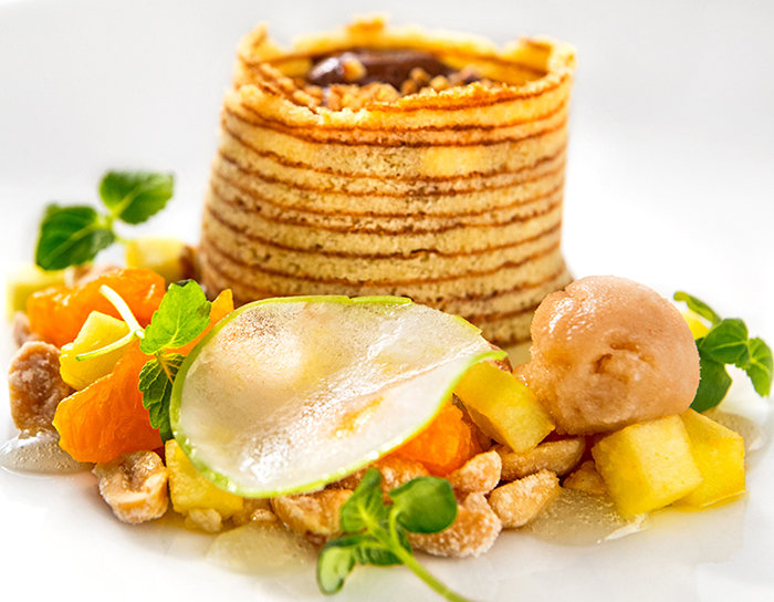 Ein Gourmet-Dessert besteht aus einem hohen, spiralförmigen Gebäckturm gefüllt mit Nüssen, umgeben von Obstwürfeln, einer dünnen Birnenscheibe, Grünzeug und einer Kugel Sorbet, alles elegant auf einem weißen Teller präsentiert.