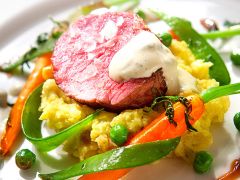 Ein Gourmetgericht mit einer Scheibe blutigem Rindfleisch, garniert mit Sahnesauce, serviert auf einem Bett aus Kartoffelpüree. Dazu hellgrüne Erbsen, sautierte Zuckerschoten und glasierte Karotten, garniert mit Kräutern.