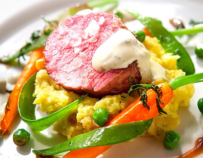Ein Gourmetgericht mit einer Scheibe blutigem Rindfleisch, garniert mit Sahnesauce, serviert auf einem Bett aus Kartoffelpüree. Dazu hellgrüne Erbsen, sautierte Zuckerschoten und glasierte Karotten, garniert mit Kräutern.