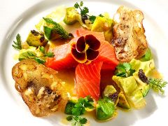 Ein Gourmetgericht mit dünn geschnittenem Lachs, garniert mit einer essbaren Blüte. Es ist umgeben von Rosenkohlblättern, Kräutern, Walnüssen und knusprigen Brotscheiben auf einem weißen Teller.