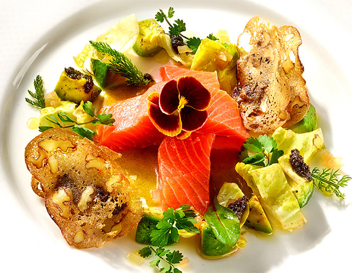 Ein Gourmetgericht mit dünn geschnittenem Lachs, garniert mit einer essbaren Blüte. Es ist umgeben von Rosenkohlblättern, Kräutern, Walnüssen und knusprigen Brotscheiben auf einem weißen Teller.