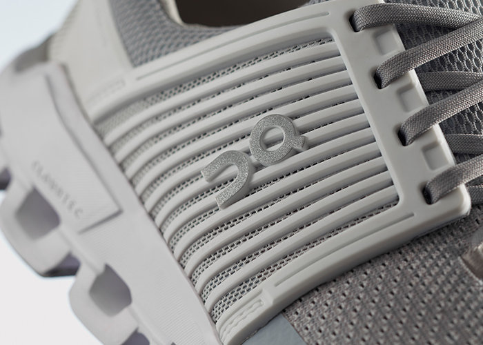 Nahaufnahme eines grauen Sportschuhs mit einem einzigartigen gerippten Design und einem Logo mit den Buchstaben „ON“. Der Schuh hat eine strukturierte Netzoberfläche und dicke, eckige Sohlenelemente, wobei die runden Schnürsenkel teilweise sichtbar sind.