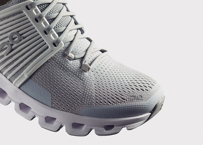 Nahaufnahme eines grauen Sportschuhs mit aufwendigen Mesh-Details und einem einzigartigen wolkenartigen Sohlendesign. Der Schuh hat hellgraue Schnürsenkel und ein dezentes Logo im Zehenbereich. Der Hintergrund ist schlicht weiß.