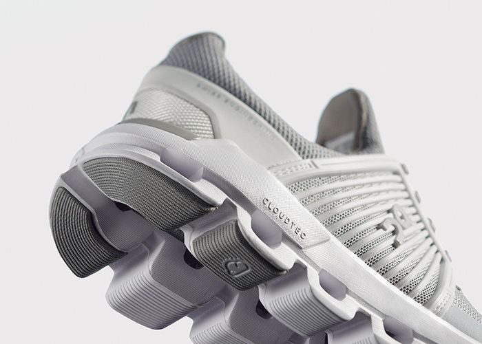 Nahaufnahme eines grau-weißen Sportschuhs mit gepolsterter Sohle und strukturiertem, segmentiertem Design. Der obere Teil besteht aus atmungsaktivem Mesh-Material und der Modellname „Cloudtec“ ist an der Seite sichtbar.