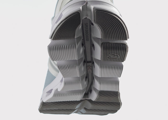 Nahaufnahme von zwei Schuhsohlen mit einem gitterartigen Muster und Rillen. Das Design ist grau und weiß gehalten, wobei zwischen den Sohlen das Wort „SHOEGOO“ zu sehen ist.