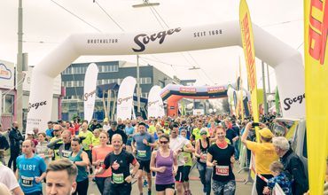 Eine große Gruppe von Läufern nimmt an einem Straßenrennen teil und läuft unter einem weißen aufblasbaren Bogen mit der Aufschrift „Sieger“ durch. Die Menge trägt verschiedene Sportkleidung und die Atmosphäre ist energiegeladen mit bunten Bannern und Zuschauern an den Seiten.
