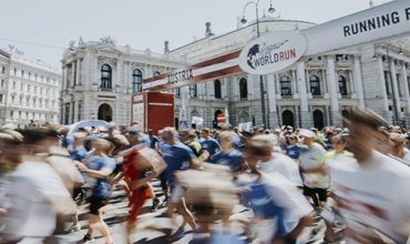 Eine große Menge Läufer nimmt an einem Marathon vor einem historischen Gebäude teil. Die Szene ist dynamisch, die Energie wird durch verschwommene Bewegungen eingefangen. Auf einem Banner über dem Lauf sind die Worte „World Run“ und „Österreich“ als Teil der Veranstaltungsbeschilderung zu sehen.