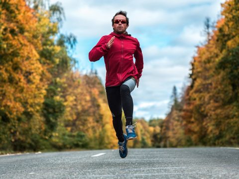 Eine Person in einer roten Jacke und Sonnenbrille sprintet auf einer Straße. Die Straße ist von Bäumen mit Herbstlaub in leuchtenden Gelb- und Orangetönen umgeben, unter einem bewölkten Himmel.