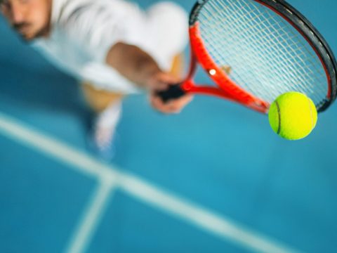 Ein Tennisspieler in einem weißen Hemd ist von oben aufgenommen, wie er mit einem roten Schläger einen leuchtend gelben Tennisball auf einem blauen Platz schlägt. Das Bild ist leicht verschwommen, wodurch die Bewegung und der Fokus auf den Ball betont werden.