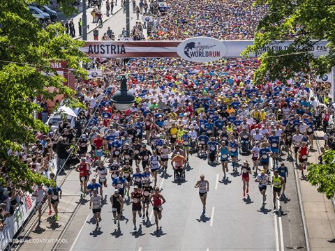 Eine große Menge Läufer nimmt unter einem Banner mit der Aufschrift „Austria“ und „World Run“ an einem Marathon teil. Die Straße ist voll mit Menschen aller Altersgruppen, einige davon auf Fahrrädern. Bäume säumen die Straße, und die Atmosphäre ist lebhaft und energiegeladen.