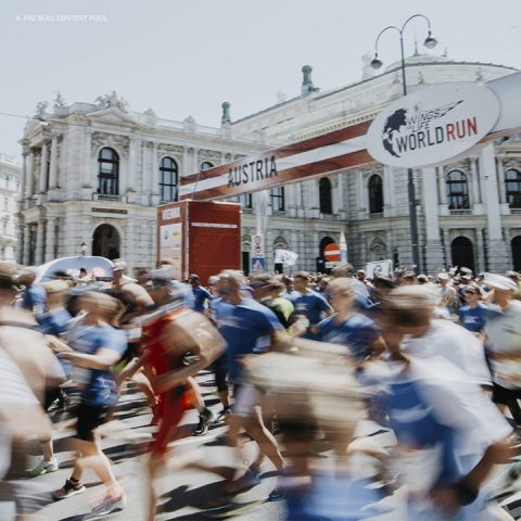 Unscharfes Bild einer großen Gruppe von Läufern, die am Wings for Life World Run vor einem historischen Gebäude in Wien, Österreich, teilnehmen. Ein Banner über ihnen weist auf die Veranstaltung hin. Die Menschen tragen Sportkleidung und scheinen in Bewegung zu sein.